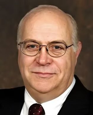 Close-up of a middle-aged man with glasses, wearing a suit and tie, smiling at the camera.