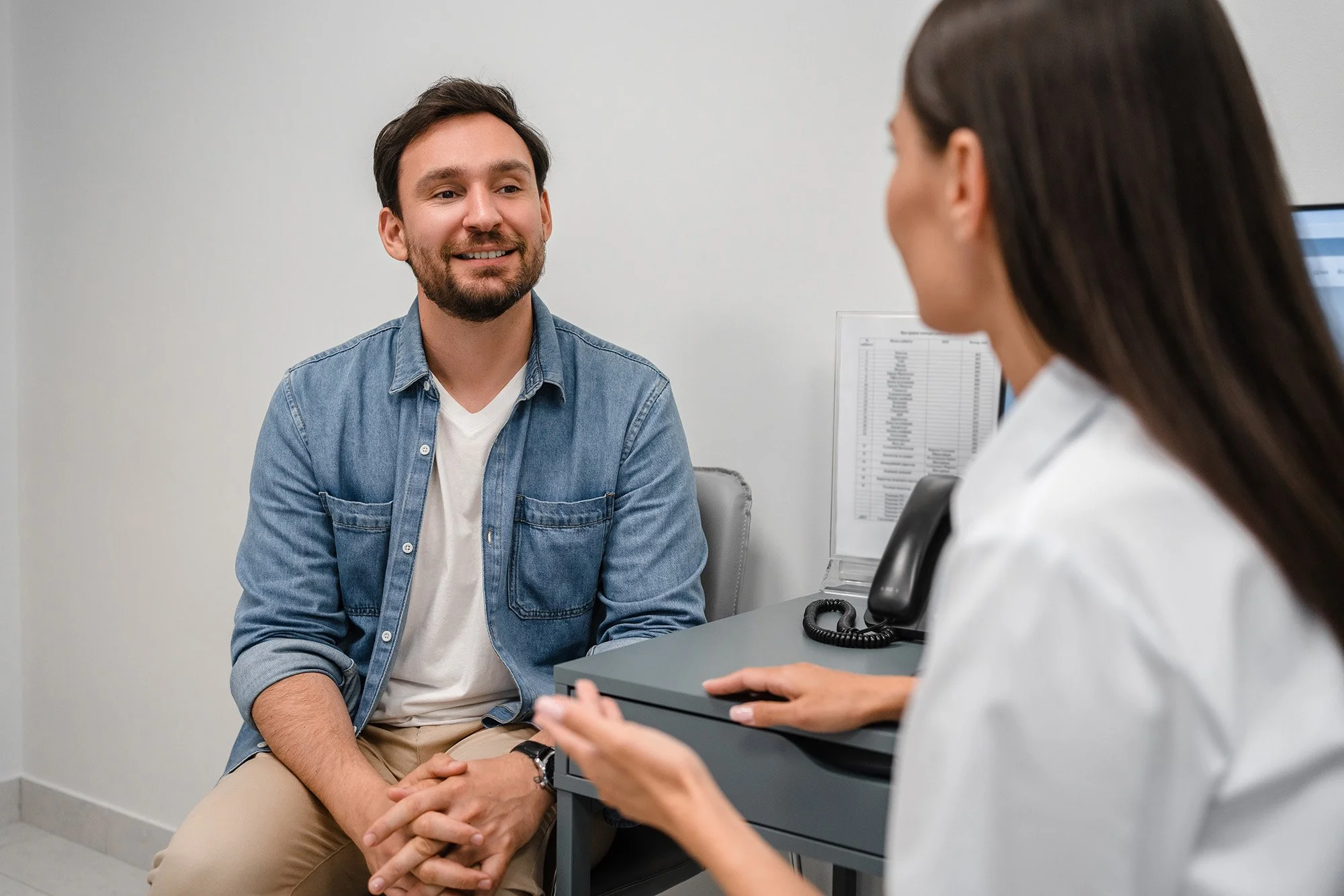 A young man with dark hair and a beard smiling while sitting in a medical office, talking to a female healthcare professional with long dark hair, who is holding a clipboard and speaking to him.