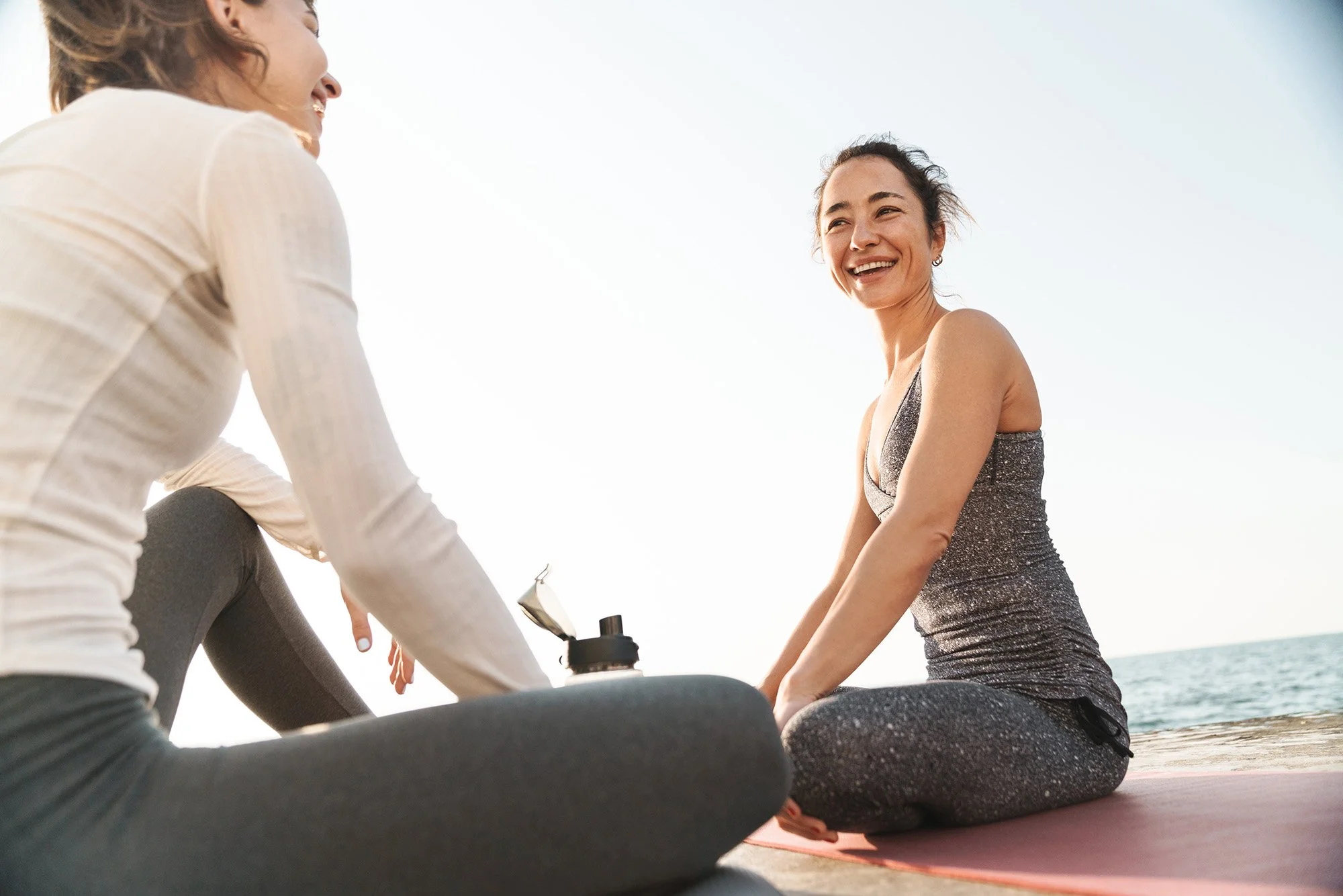 Two women practicing yoga outdoors near the ocean, sitting on yoga mats during sunset, smiling and engaging in a relaxing exercise.
