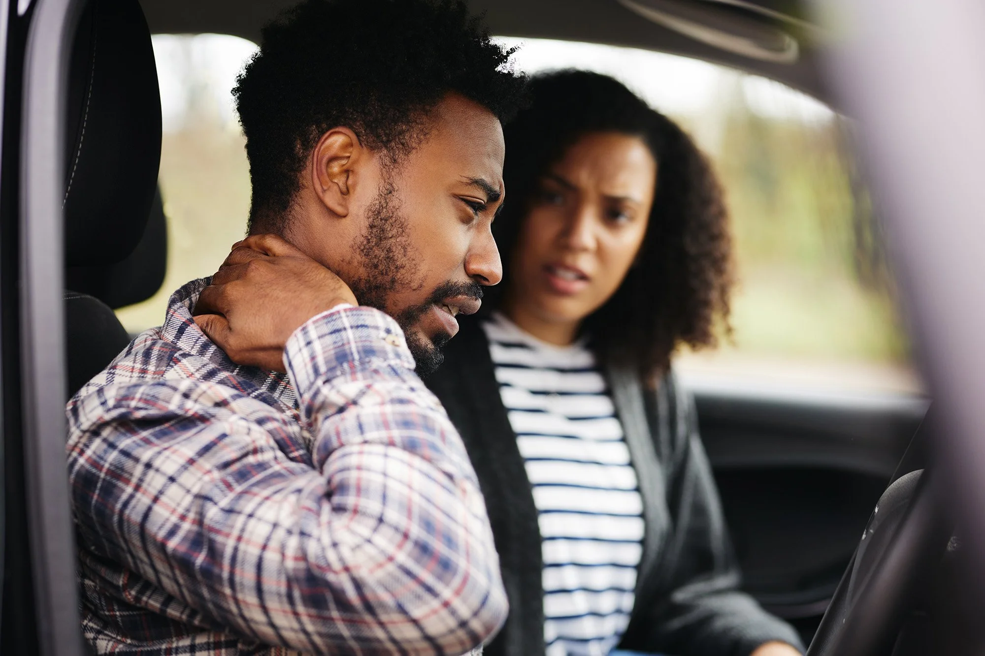 A man with a beard and curly hair sitting in a car, holding his neck with a worried expression, and a woman with curly hair sitting beside him, looking at him with concern.