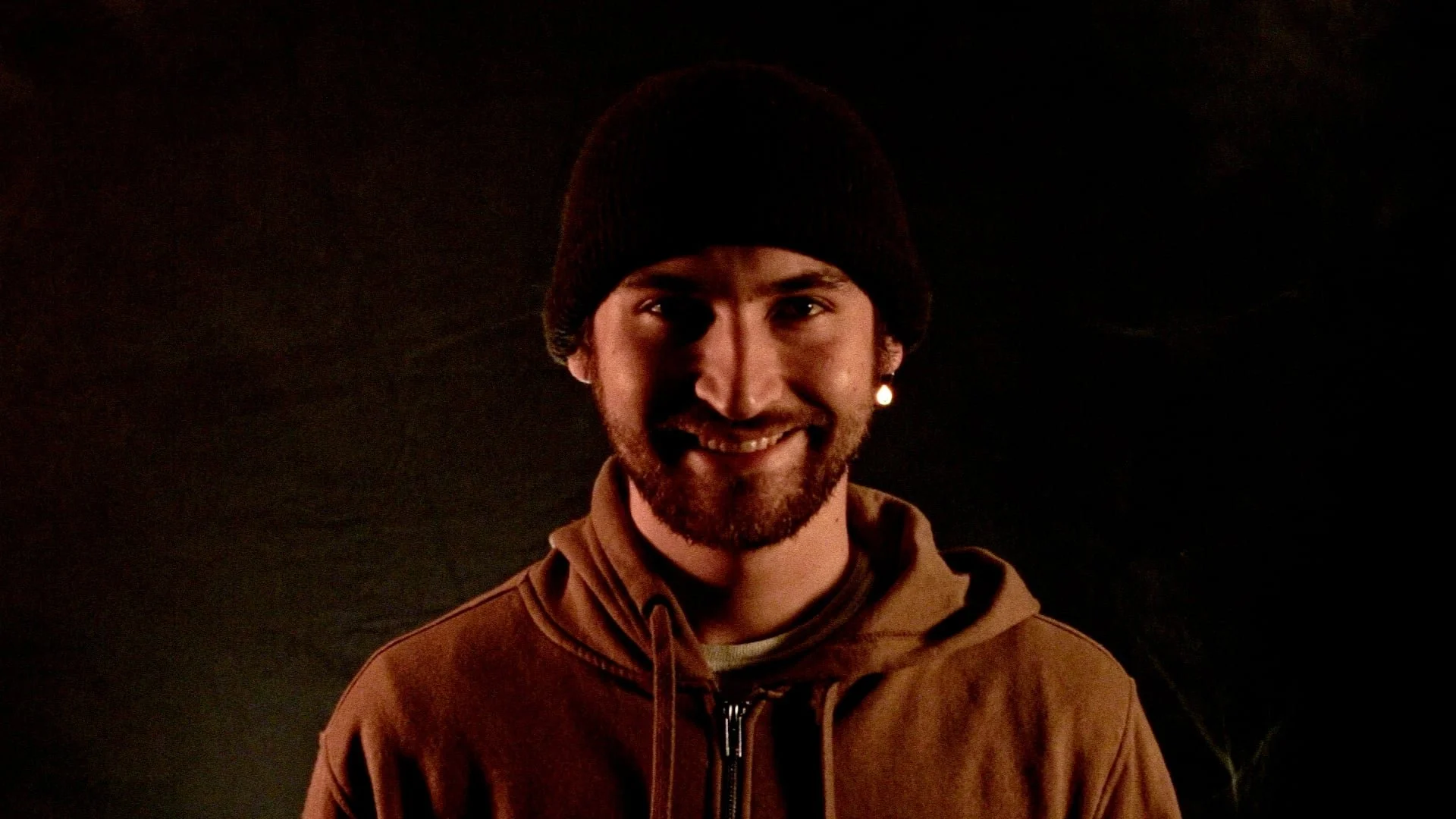 A smiling man with a beard wearing a black beanie and brown hoodie against a dark background.