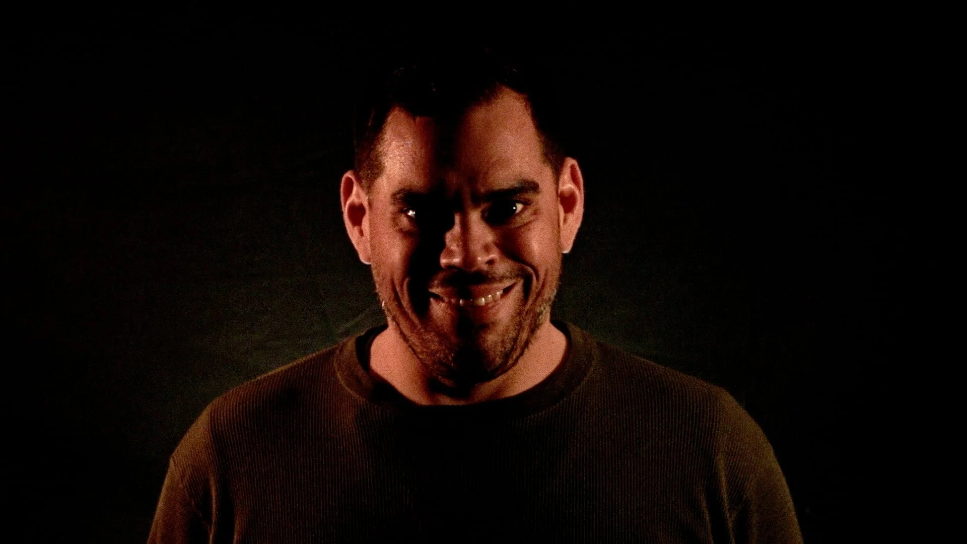 A man with short dark hair and a beard, making a mischievous smile against a dark background.