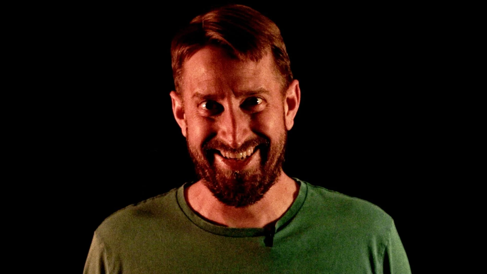A man with red hair, a beard, and a green shirt is smiling mischievously against a black background.