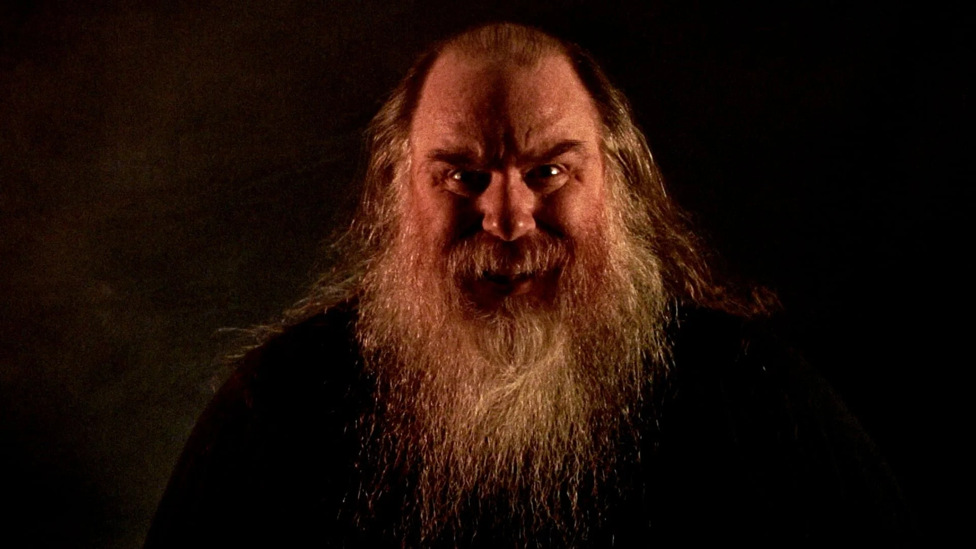 Man with long, gray beard and hair, making an angry or intense facial expression, against a dark background.