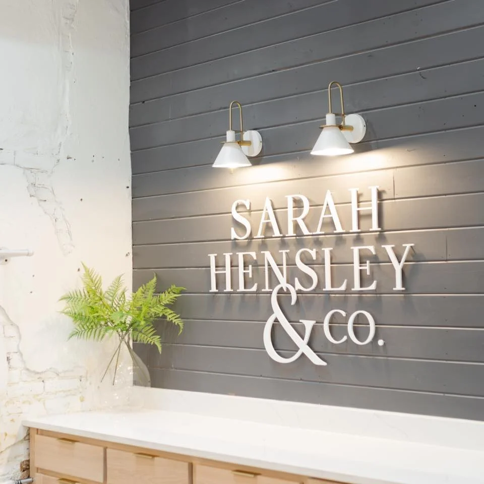 Interior of a boutique or office with a gray wooden wall, illuminated signs reading 'Sarah Hensley & Co.' and two wall-mounted lamps, a white countertop, and a potted plant.