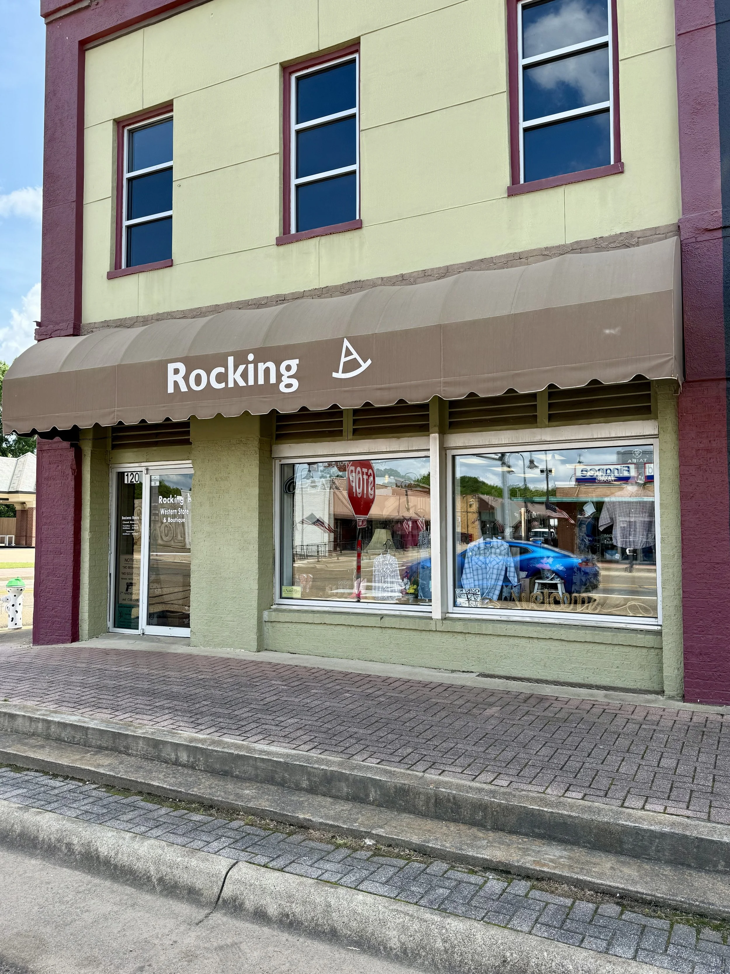 Storefront with a brown awning that says 'Rocking' and a small sailboat icon. The building is painted in pale yellow and burgundy, with three windows above the awning. The storefront has a large glass window displaying mannequins dressed in clothing and a glass door.