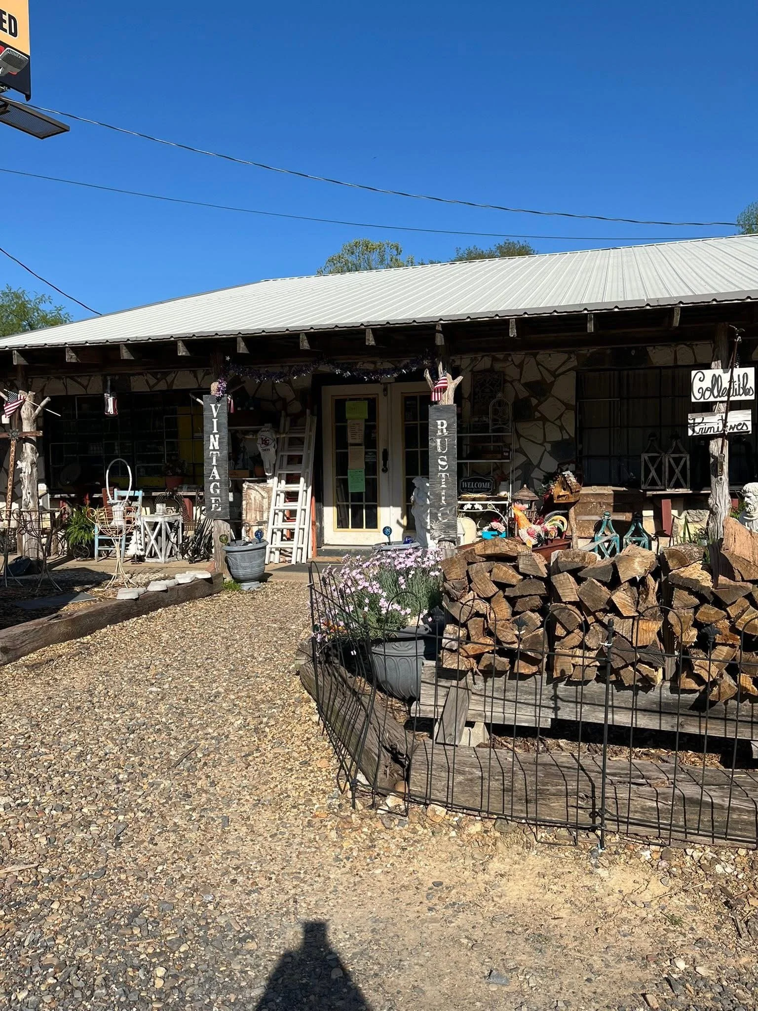 Front view of a rustic vintage shop with a gravel pathway leading to the entrance, decorated with plants and stacked firewood.
