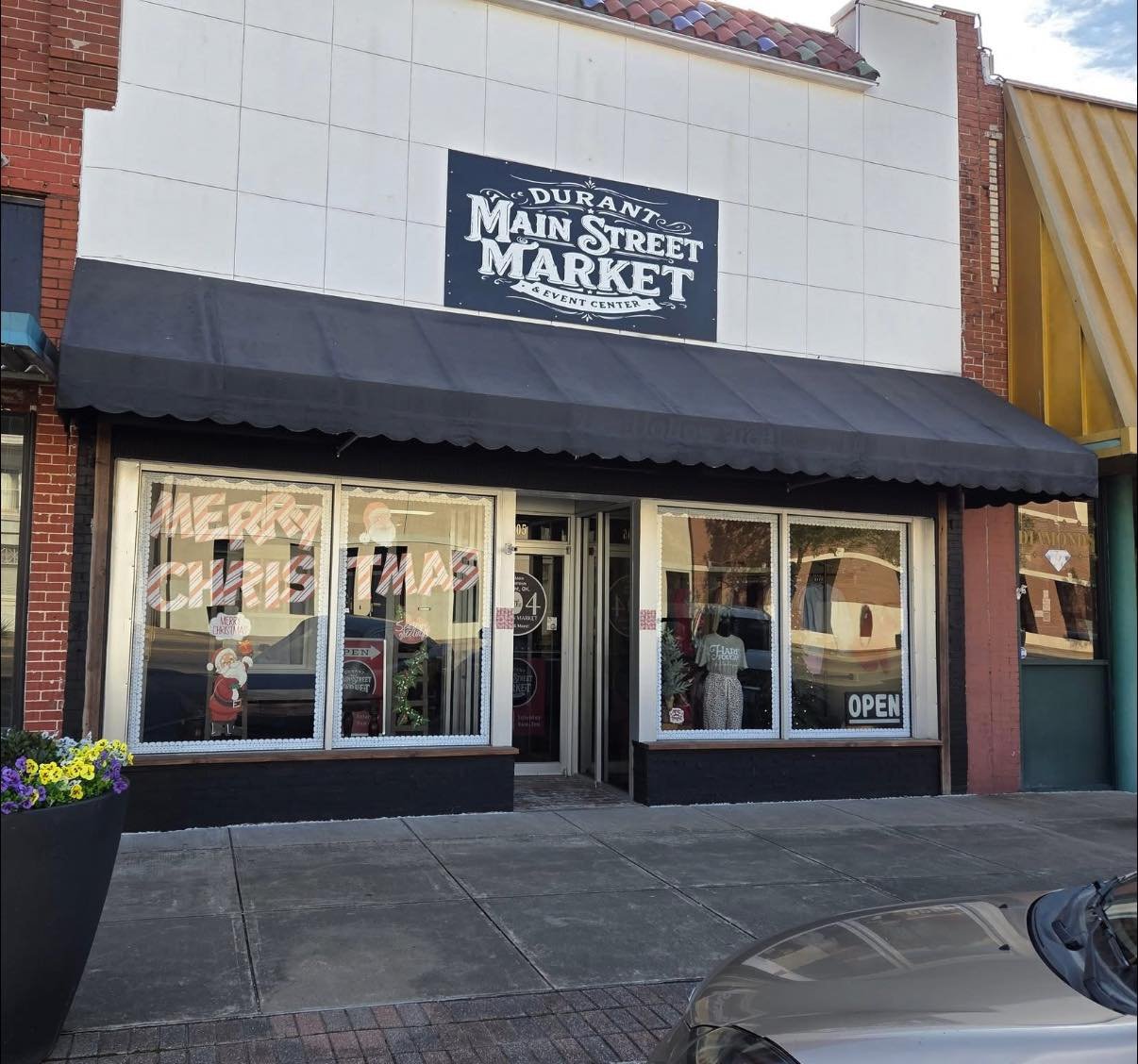  storefront with a black awning and large window displays decorated for Christmas, located at Durant Main Street Market, aevant center, with a sign above. Christmas decorations and an 'OPEN' sign are visible inside the store.