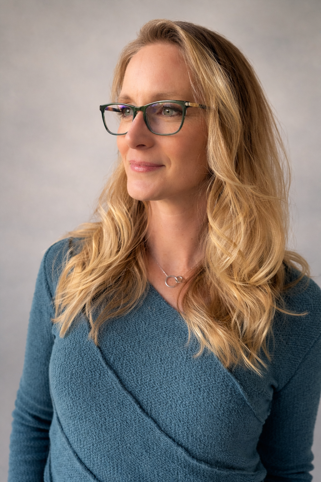 A woman with blonde, wavy hair wearing glasses and a blue textured top, smiling slightly and looking off to the side against a plain, light-colored background.