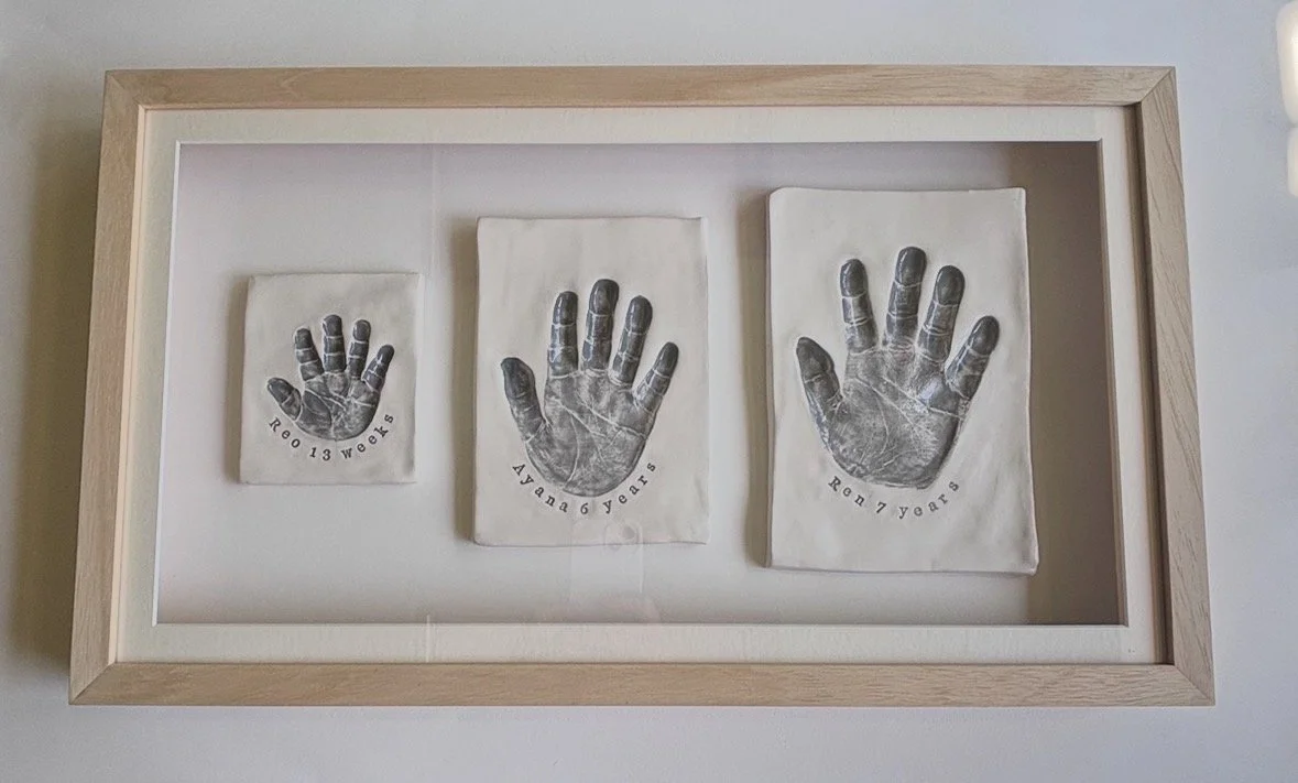 Framed display with three glazed paperweight pieces, each featuring a handprint and stamped with age: 13 weeks, 6 years, and 7 years.