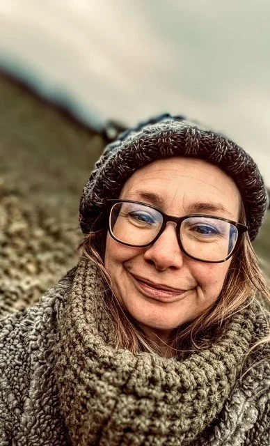 Close-up of a smiling woman wearing glasses, who is the owner of Mini Potters. She is wearing a knit hat, and a scarf outdoors, with a blurred natural landscape in the background.