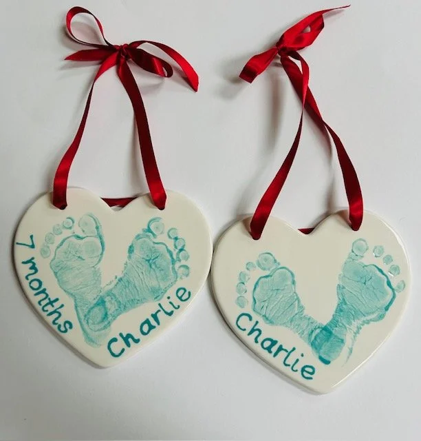 Two heart-shaped ceramic ornaments featuring baby footprints, one labeled '7 months Charlie' and the other simply 'Charlie,' with red ribbons for hanging.