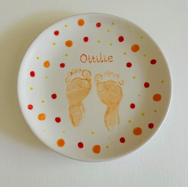Decorative white plate with orange, red, and yellow dots, featuring the name Ottilie and two sketches of baby feet.