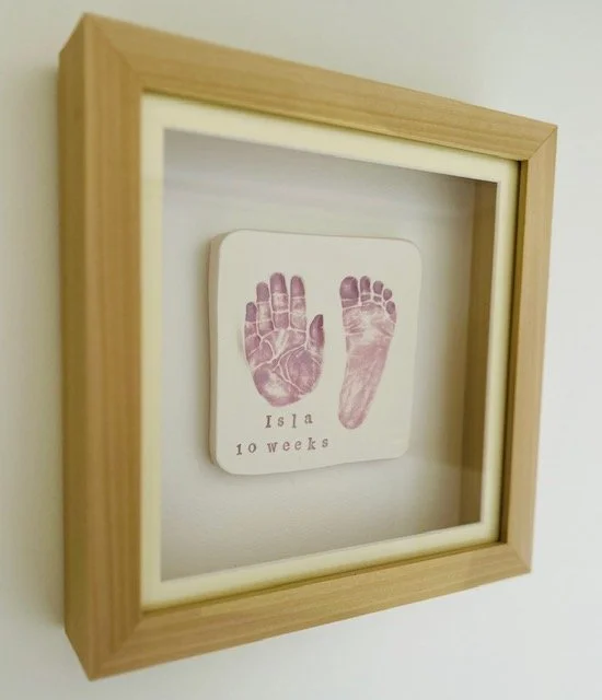 Wooden frame holds a white glazed paperweight with purple baby handprint and footprint, labelled 'Isla 10 weeks'.