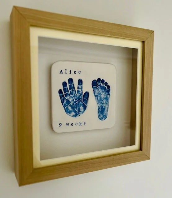 A framed display of blue handprints and footprints of a baby girl named Alice at 9 weeks old, mounted on a white background.
