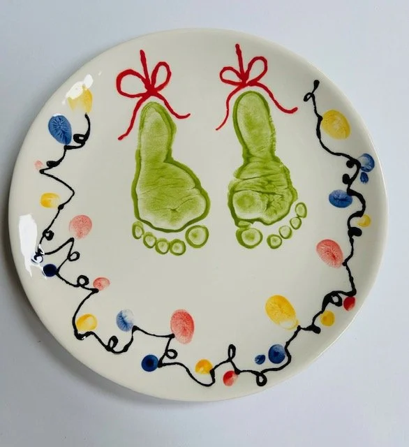 Plate decorated with colourful string lights and two green baby footprints with red ribbons tied at the top for a Christmas keepsake.