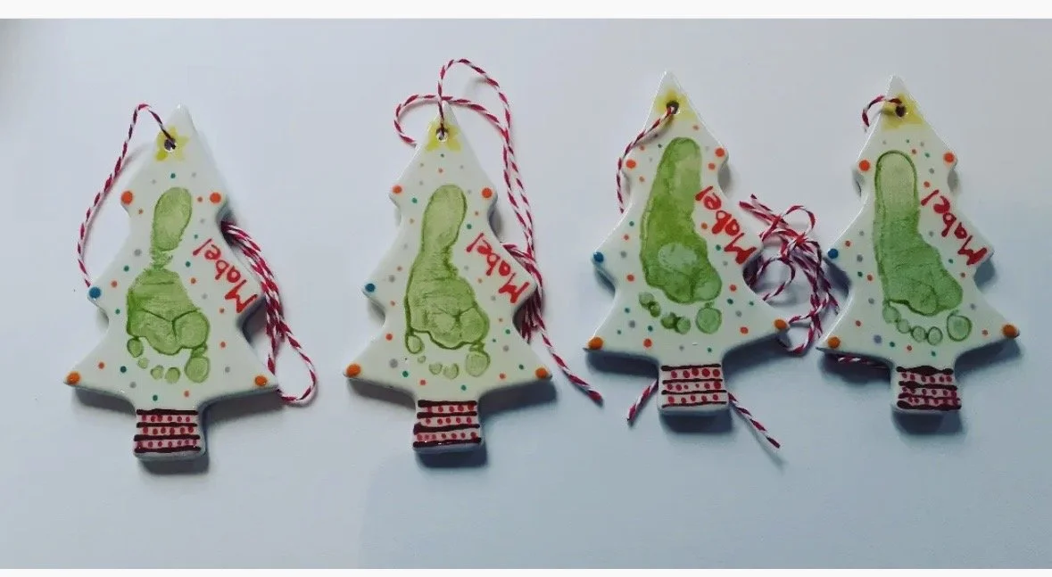 Four decorated Christmas tree-shaped decorations with green footprints, red and green polka dots, and striped red and white bases. Each decoration has a red and white string attached for hanging, with the name on each one.