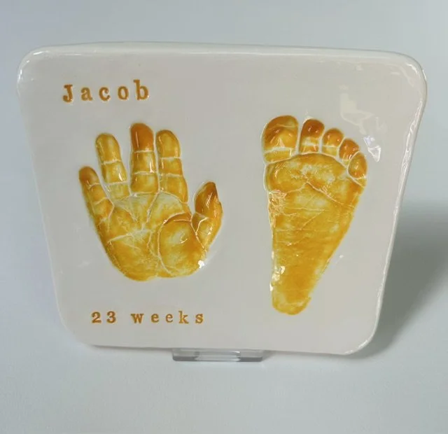 A small glazed paperweight plaque with orange baby hand and foot print, labelled 'Jacob 23 weeks.'