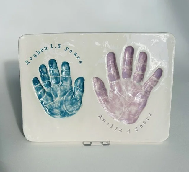 A white glazed paperweight with blue and pink handprints, celebrating the 1.5 years and 4 years of two children, Reuben and Amelia.