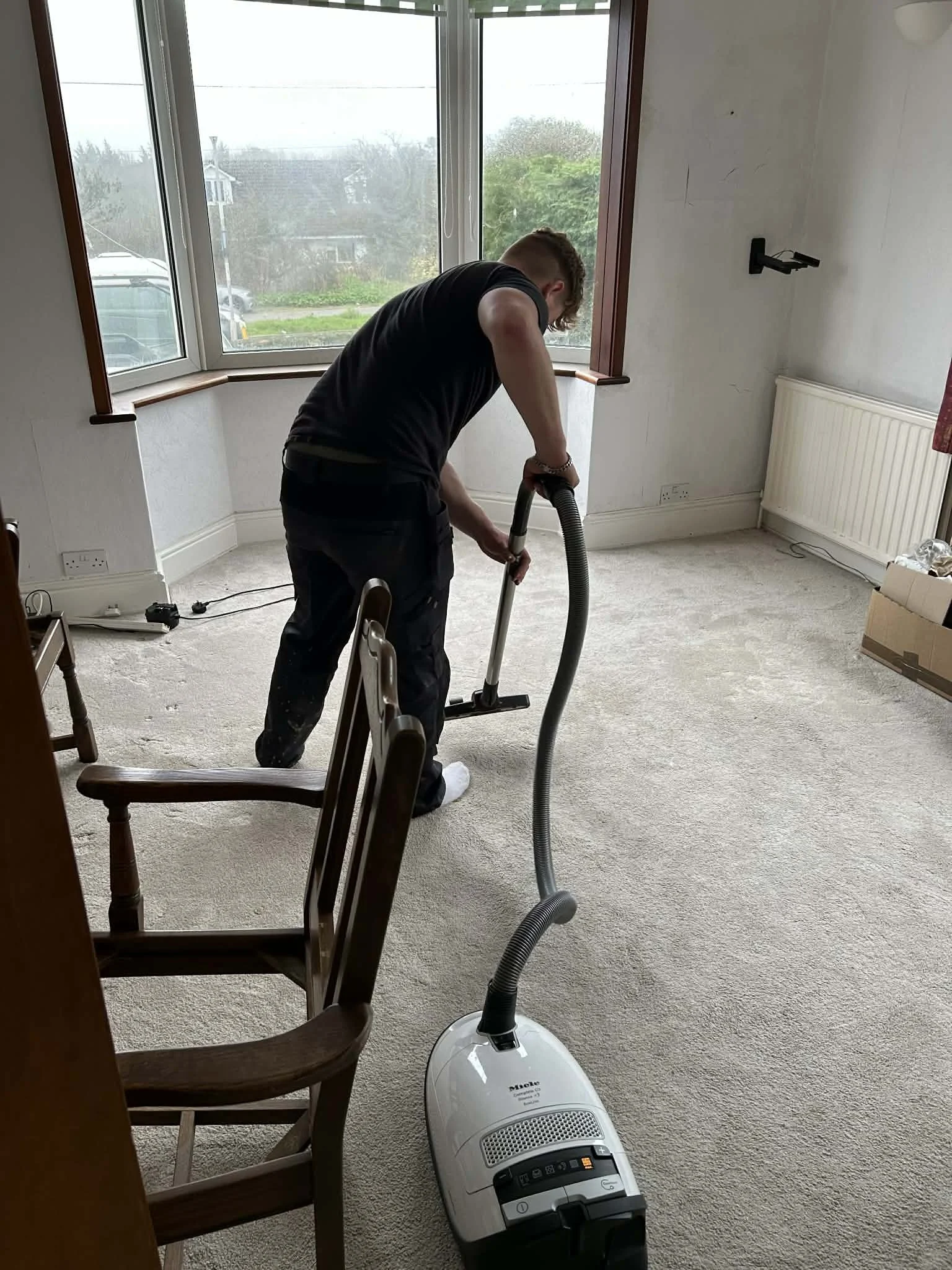 Person vacuuming beige carpet in a room with large window and white walls.