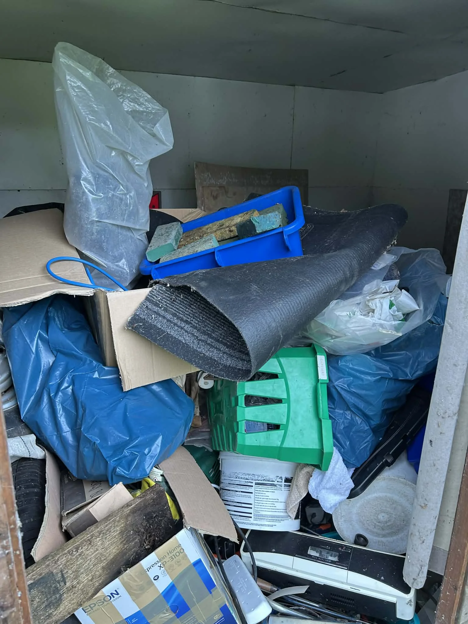 Overflowing trash and recycling in a dumpster, including cardboard, plastic bags, a green crate, and miscellaneous debris.