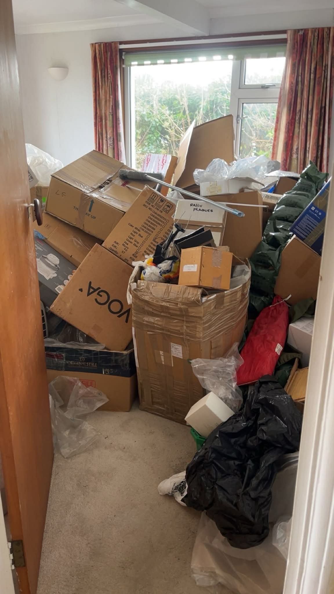 A cluttered room filled with cardboard boxes, plastic bags, and various items with a window and curtains in the background.