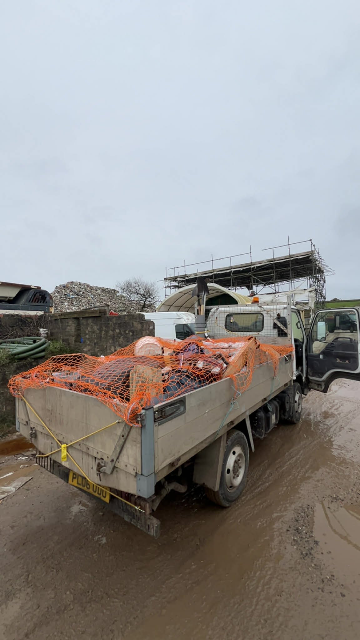 A work truck parked on a muddy construction site, loaded with tools and supplies covered with an orange safety net, with a scaffold and construction materials in the background.