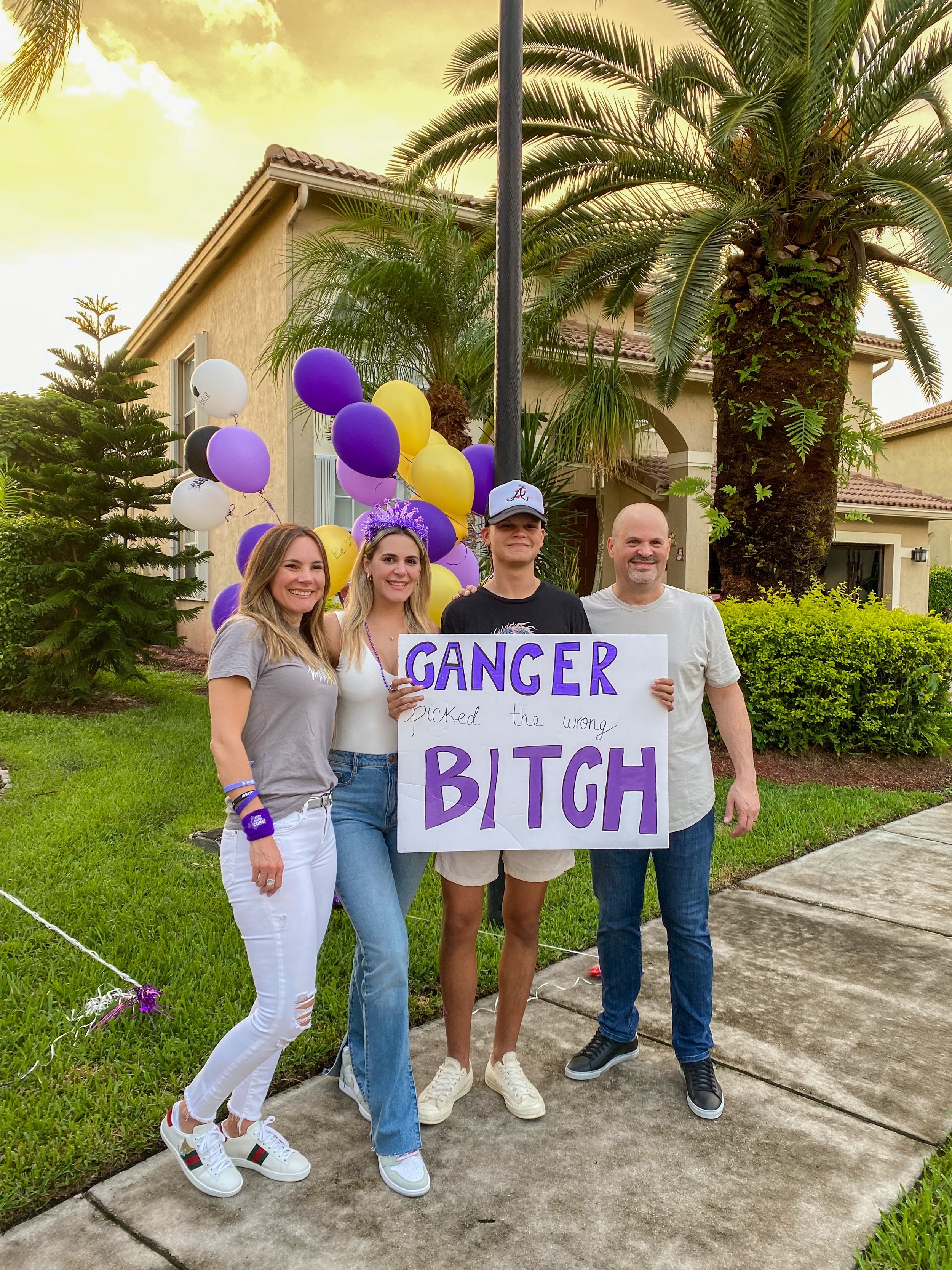 Group of four people standing outside on a sidewalk, holding a sign that says 'CANCER PICKED THE WRONG BITCH', with purple, yellow, and white balloons in the background, in front of a house with palm trees and greenery.
