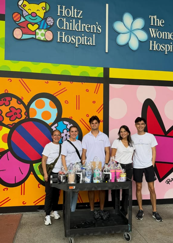 Five young people standing behind a table with gift baskets in front of colorful artwork on a hospital wall, with signs for Holtz Children's Hospital and The Women's Hospital.