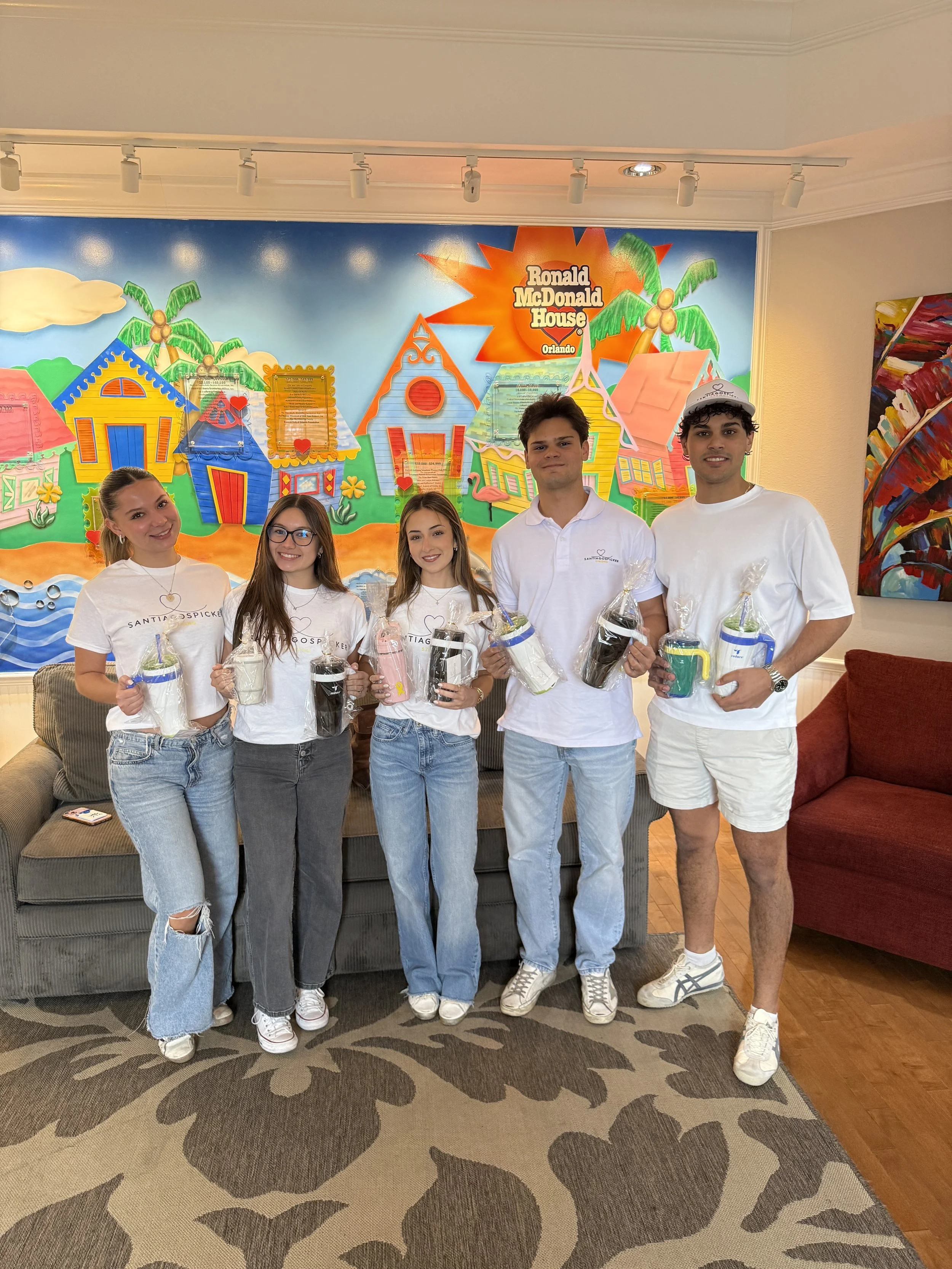 Five young people standing in front of a colorful mural at Ronald McDonald House, holding wrapped gifts or care packages.