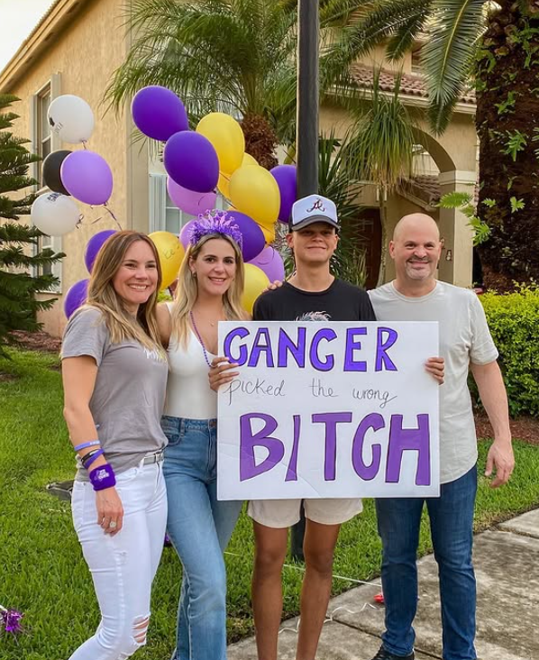 Four people standing outdoors in front of purple and yellow balloons and palm trees, holding a sign that reads "CANCER picked the wrong BITCH".