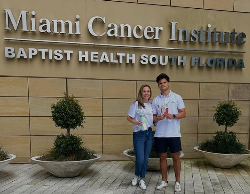 Two young adults, a woman and a man, standing in front of the Miami Cancer Institute sign at Baptist Health South Florida. They are smiling and holding trophies or awards.