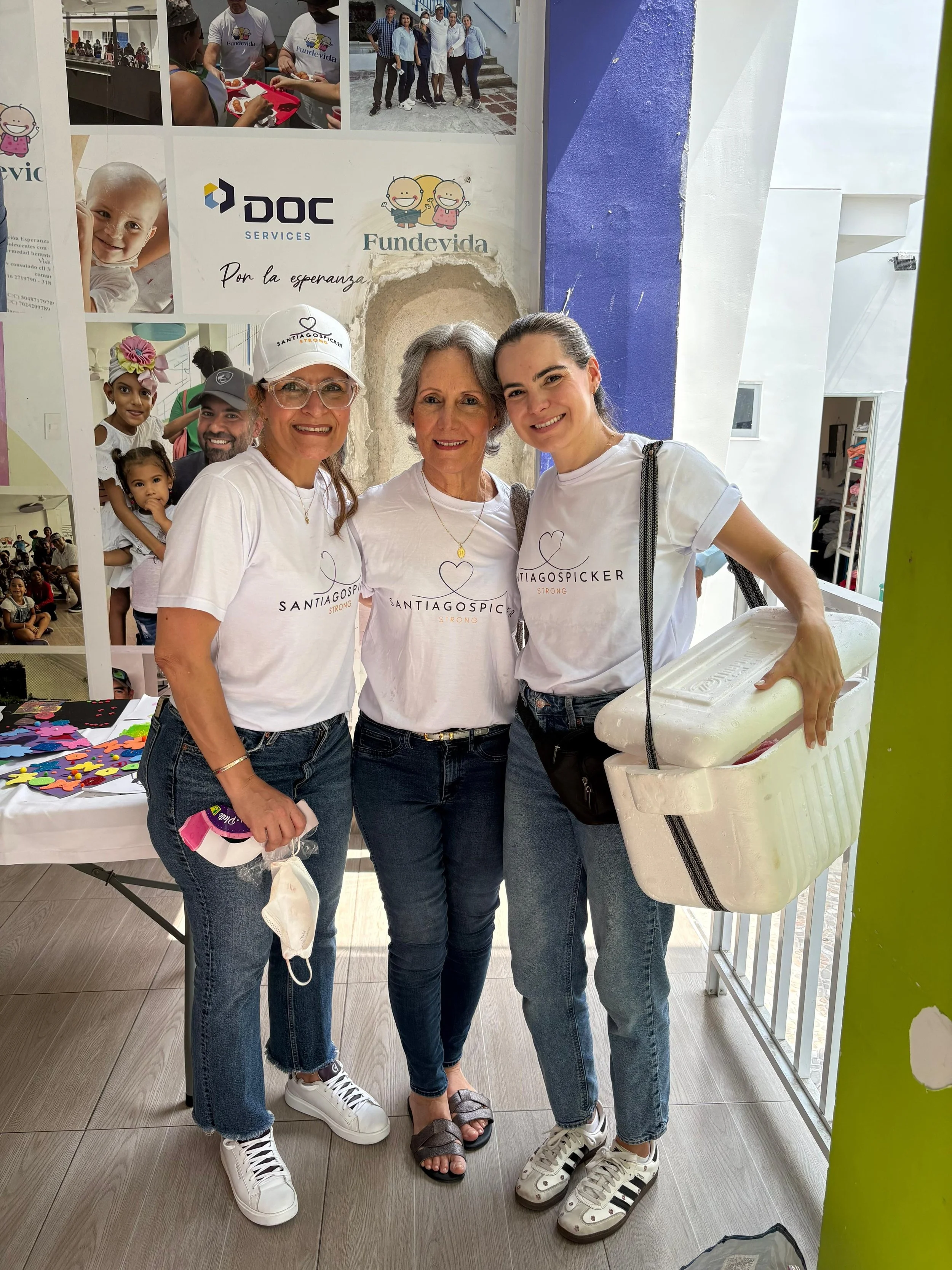 Three women wearing white T-shirts with 'SANTIAGOSPIKER' logo, smiling and posing together at a community event, with a promotional poster and children in the background.