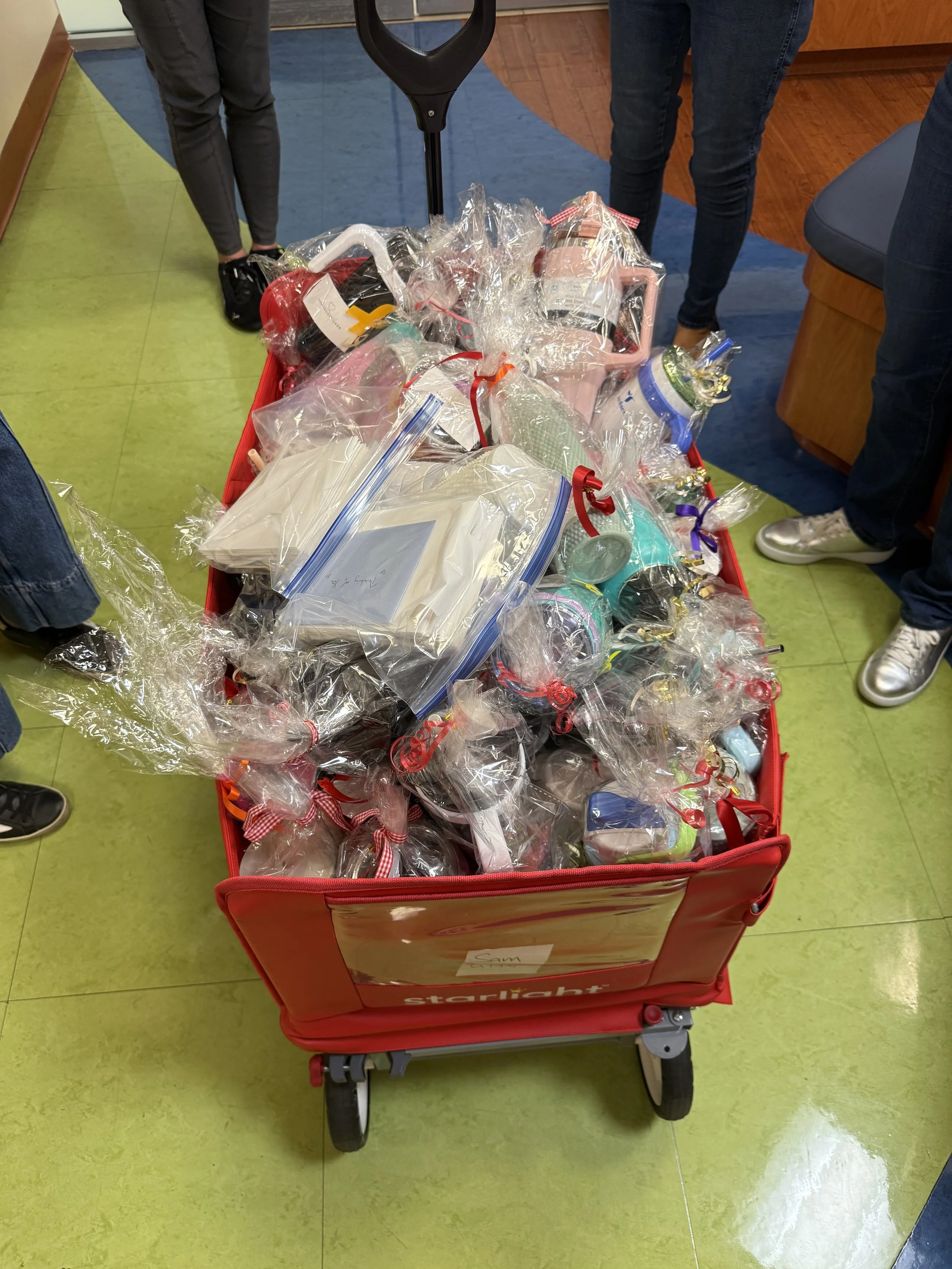 A red shopping cart filled with wrapped gifts and various items, with people standing around it in a room with green and blue tiled floors.