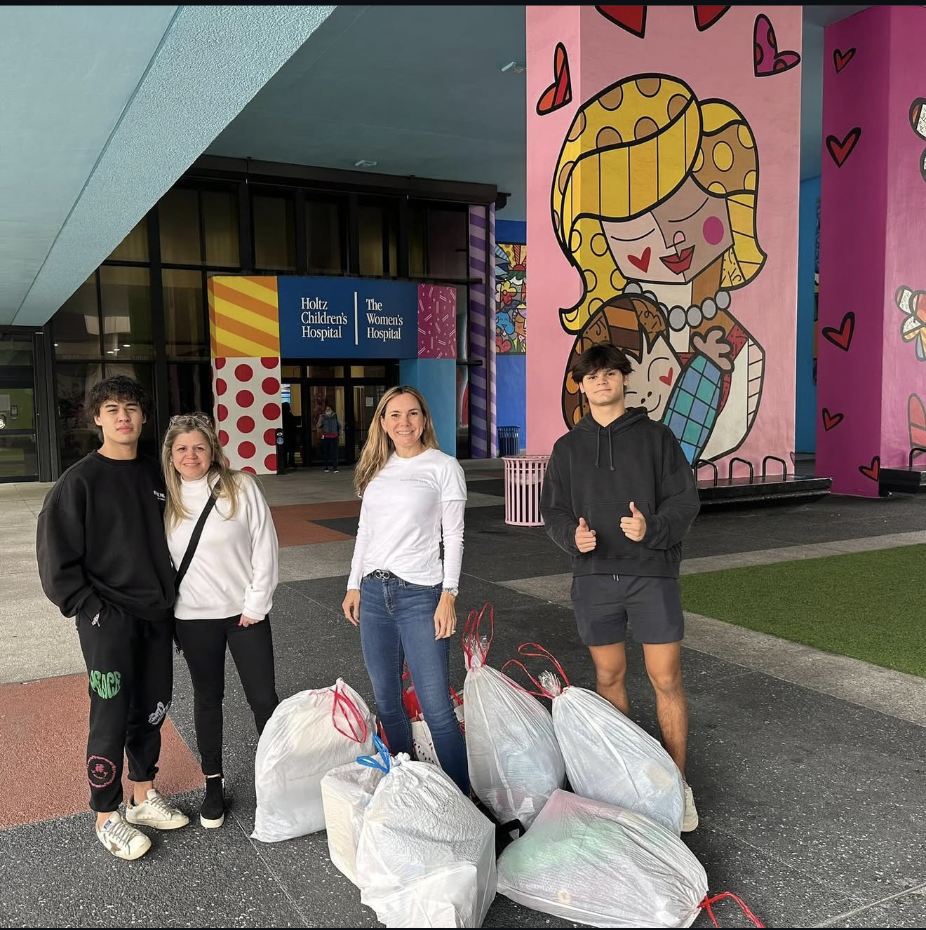 Group of four people standing outside the entrance of Holtz Children's Hospital and The Women's Hospital, holding trash bags for a cleanup.