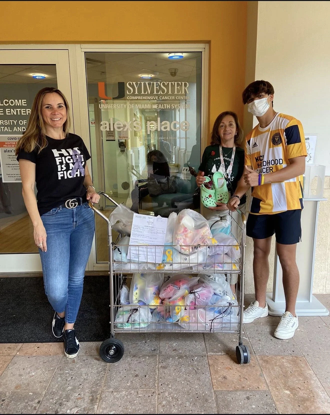 Three people standing outside a hospital entrance with a shopping cart filled with bags of goods or donations.
