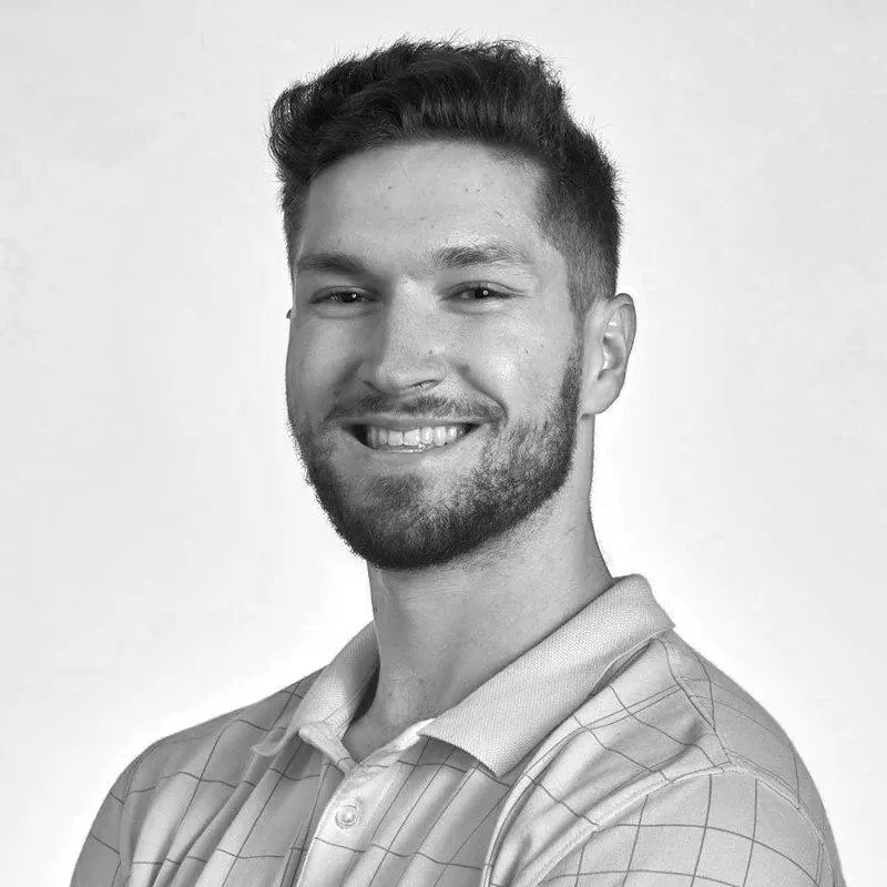 Black and white portrait of a young man with a beard and short hair, smiling, wearing a collared shirt with a checkered pattern.