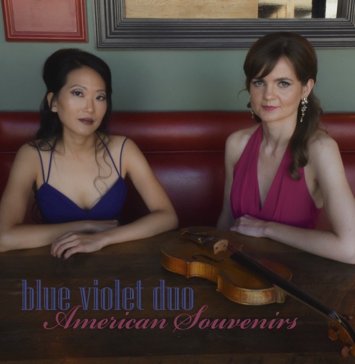 Two women sitting at a table with a violin on it, inside a restaurant or cafe, with a mirror and framed pictures in the background. The woman on the left has dark hair and is wearing a blue dress, and the woman on the right has brown hair and is wearing a pink dress. The text on the image reads 'blue violet duo' and 'American Souvenirs'.