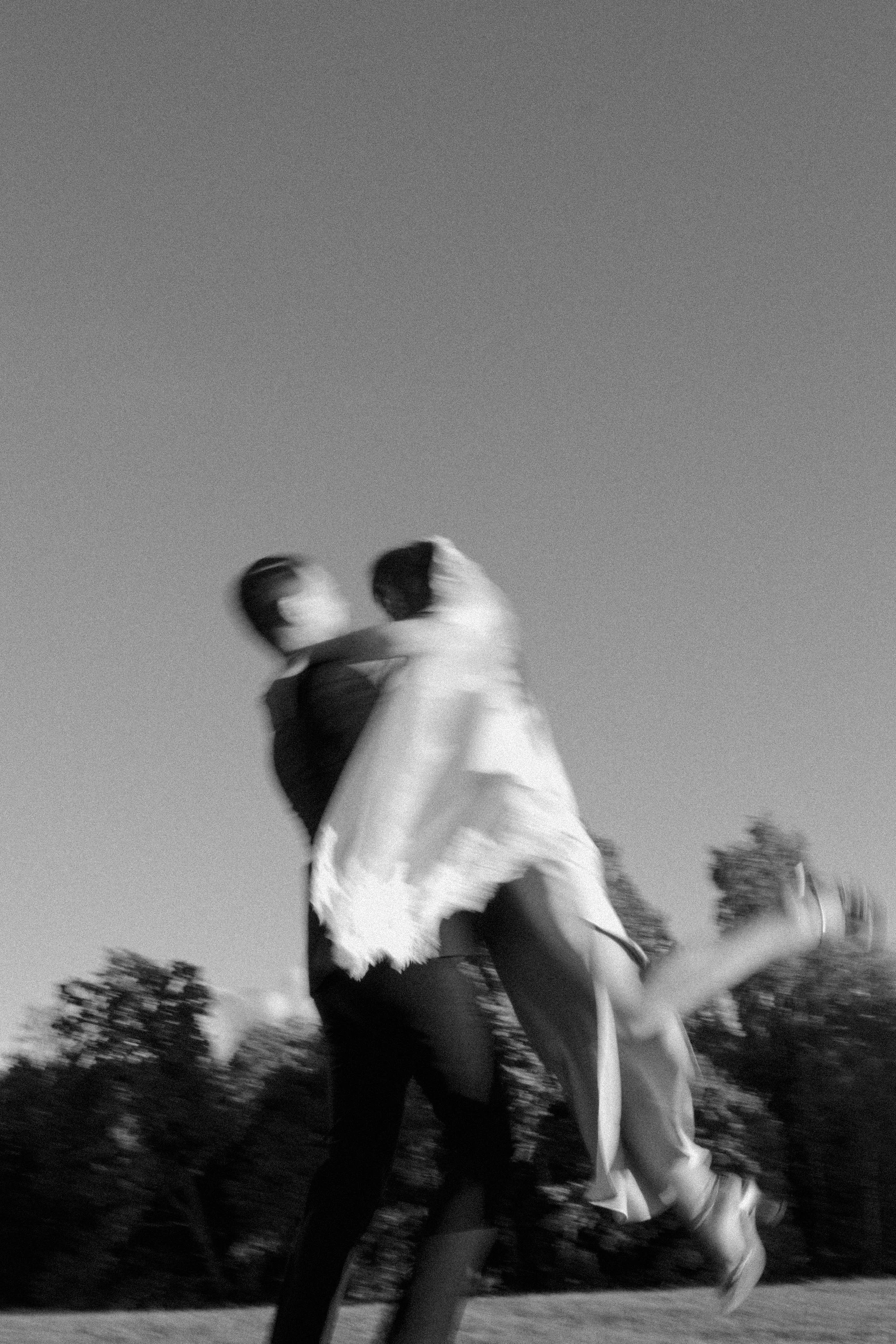 A black and white photo of a couple dancing outdoors in a field with trees in the background, motion blurred.