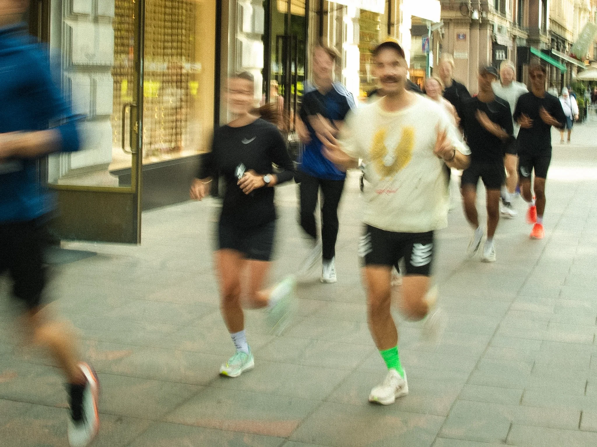 A group of runners participating in a race on a city street, wearing athletic clothing and running shoes, with buildings and pedestrians visible in the background.