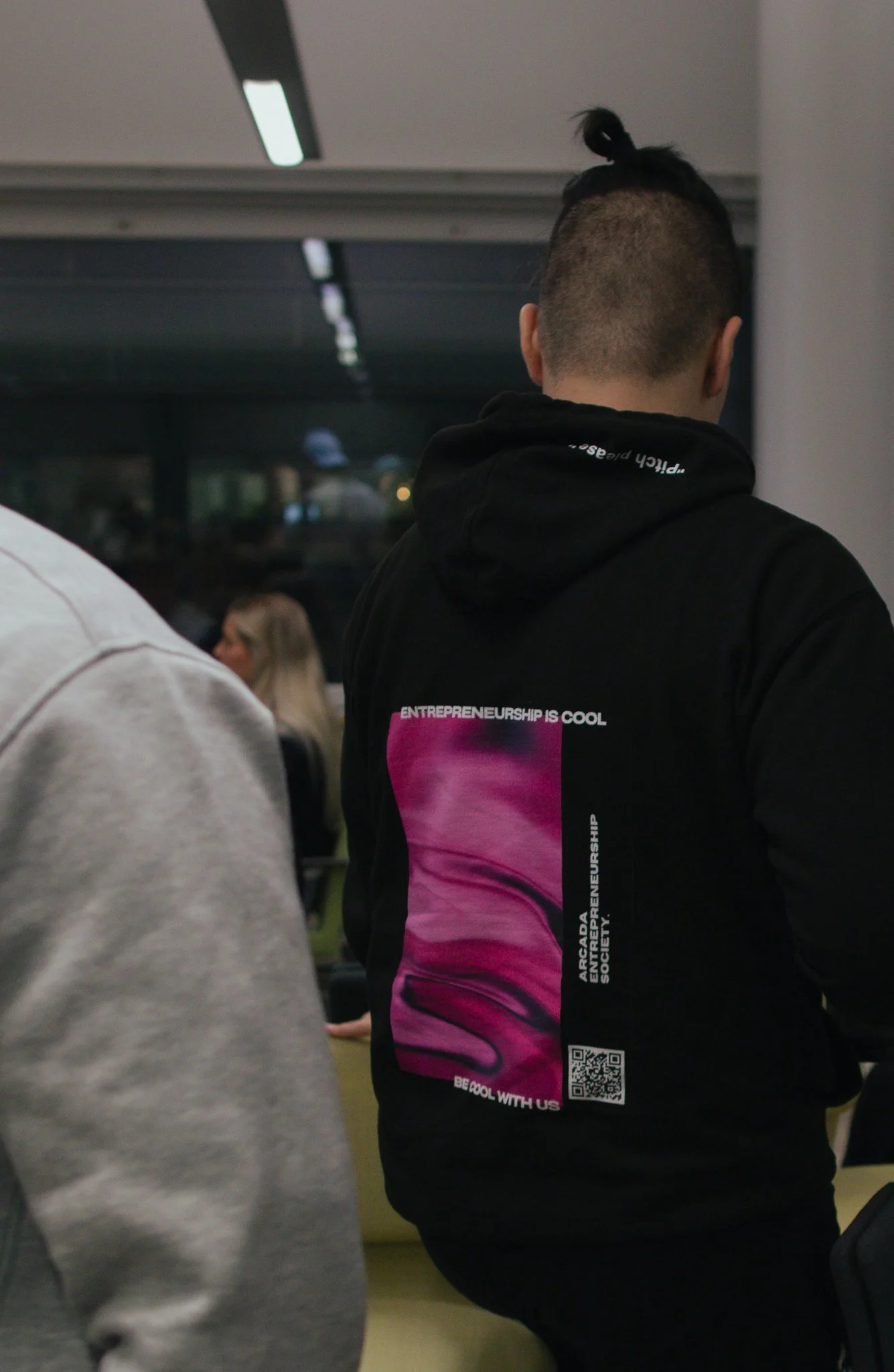 A person wearing a black hoodie with colorful graphic design on the back, with words related to entrepreneurship, standing indoors with other people seated in the background.