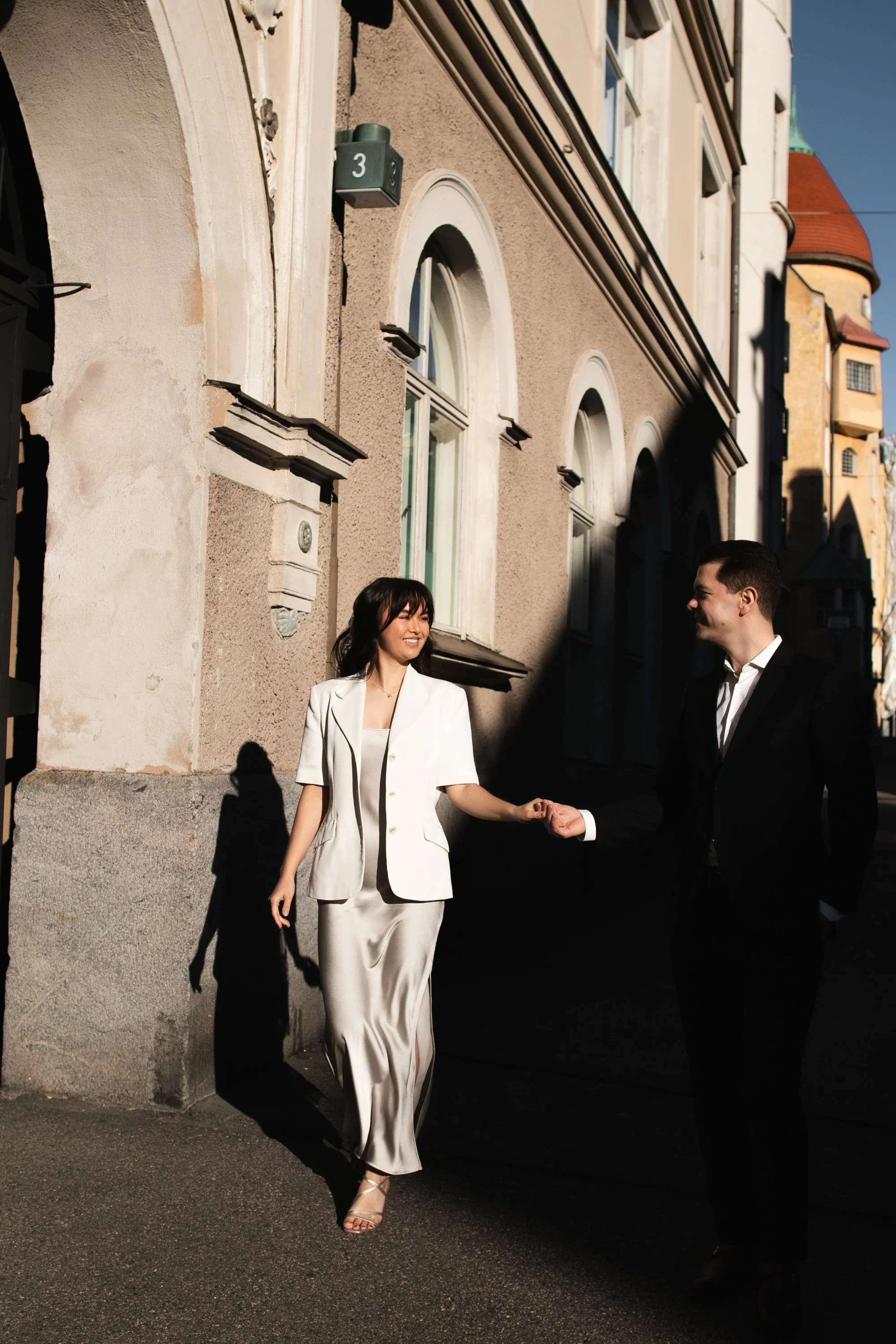 A woman in a white suit and a man in a black suit holding hands on a city street, with shadows cast on the building behind them.