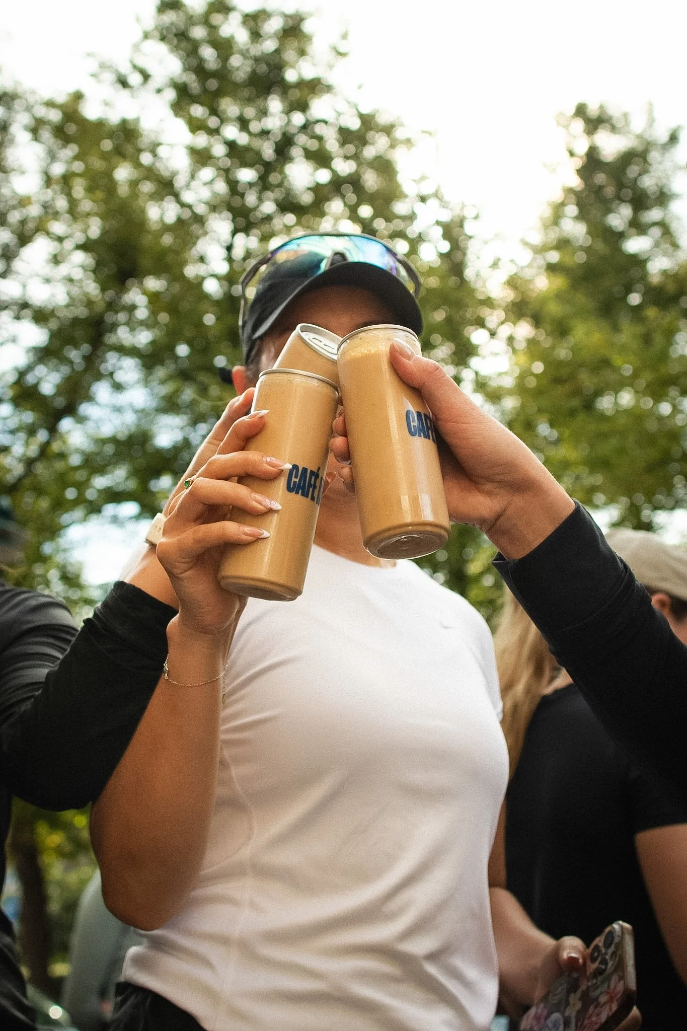 Person wearing a white shirt and black sleeves holding four cans of coffee in a park with green trees in the background.