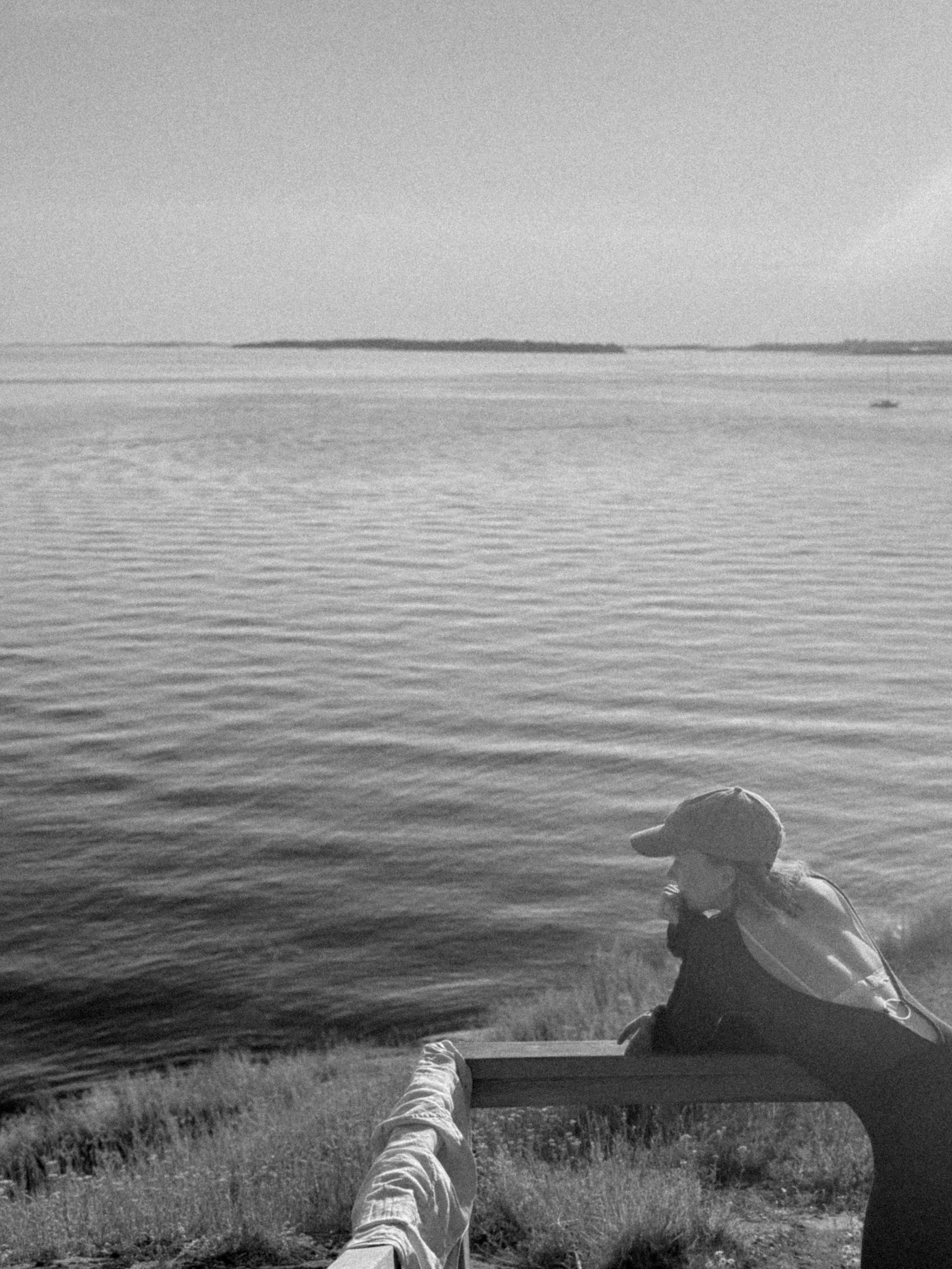 Person sitting on a bench near a body of water, looking at the water, with another person standing nearby, in black and white