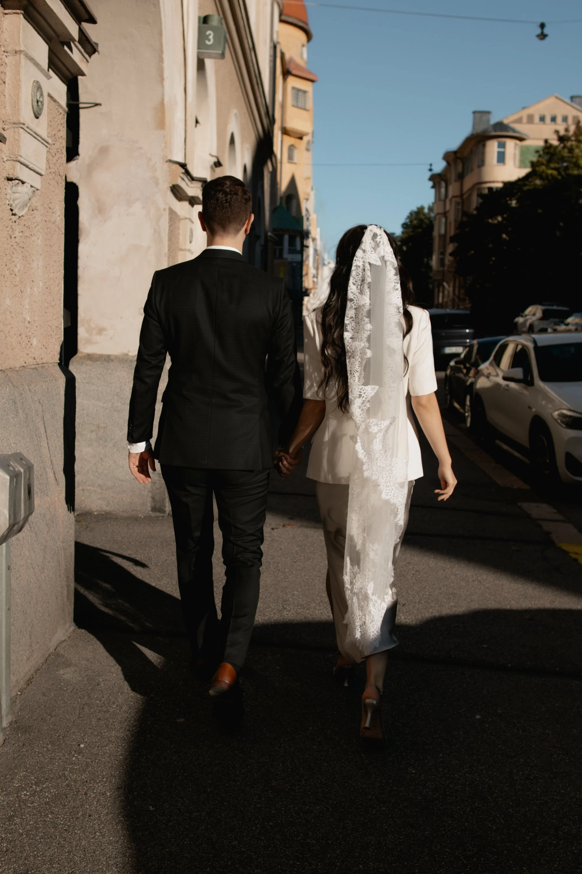 A couple walking hand in hand down a city sidewalk during daytime. The groom is wearing a black suit and brown shoes, while the bride is dressed in a light-colored outfit with a lace veil, and high heels. They are walking away from the camera, and th