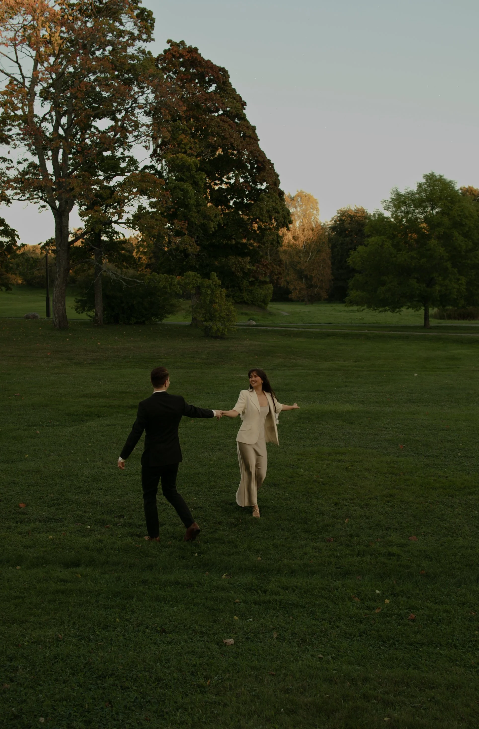 A man in a black suit and a woman in a beige suit are holding hands and dancing outdoors in a park during dusk, with trees and a grassy field in the background.
