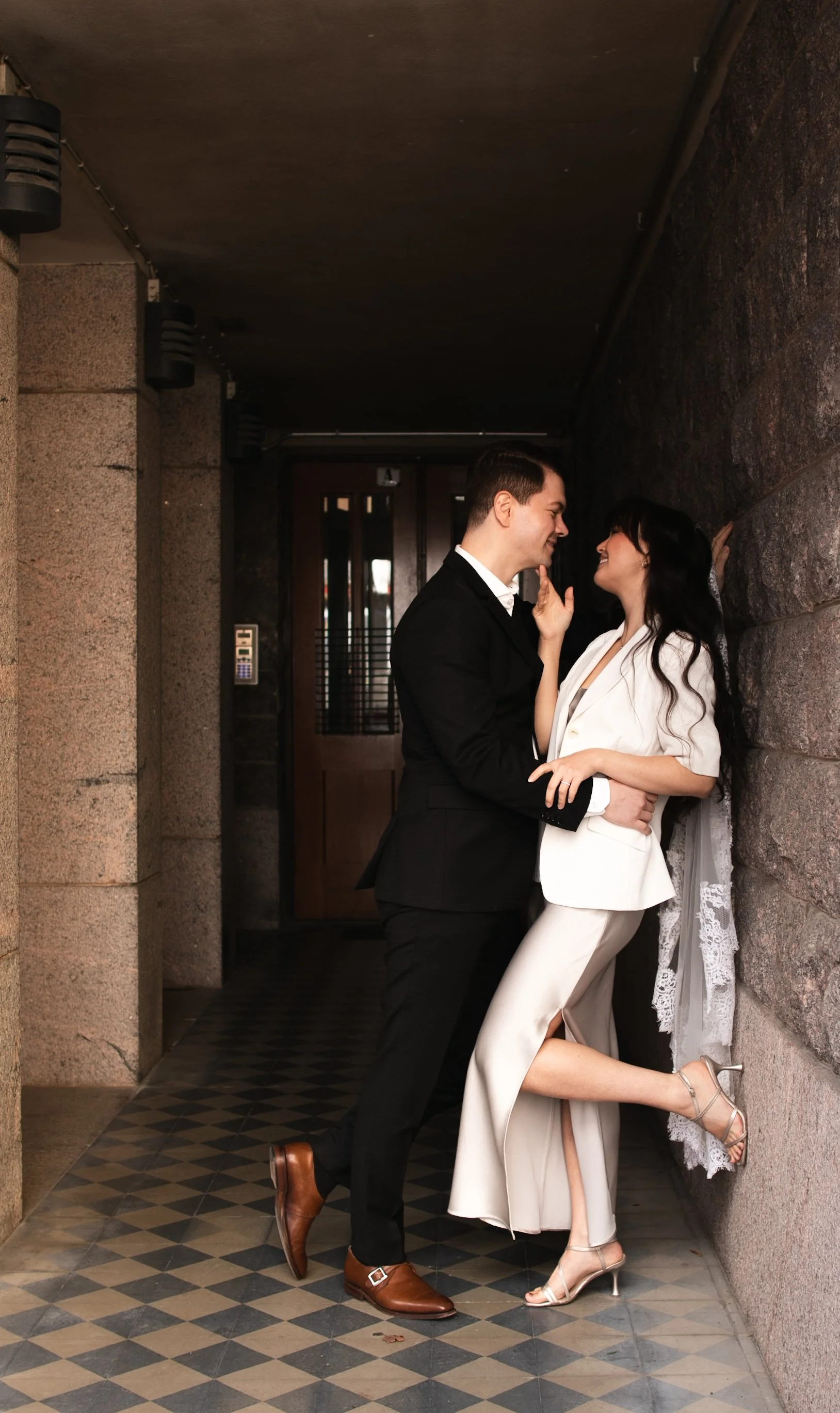 A stylish couple in formal attire, smiling and close together, standing in a dimly lit hallway with textured walls and a checkered floor.