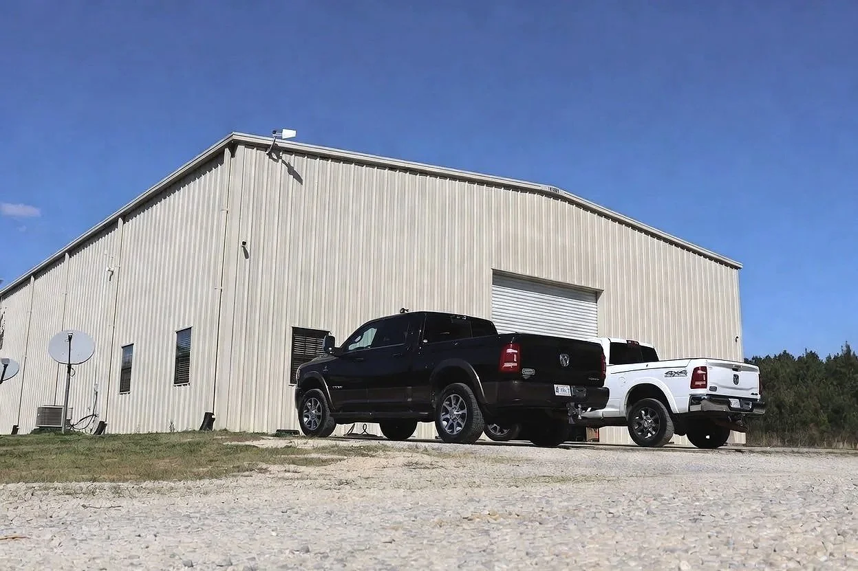 A beige metal building with a few small windows and satellite dishes, two pickup trucks parked in front, and a blue sky with some clouds.