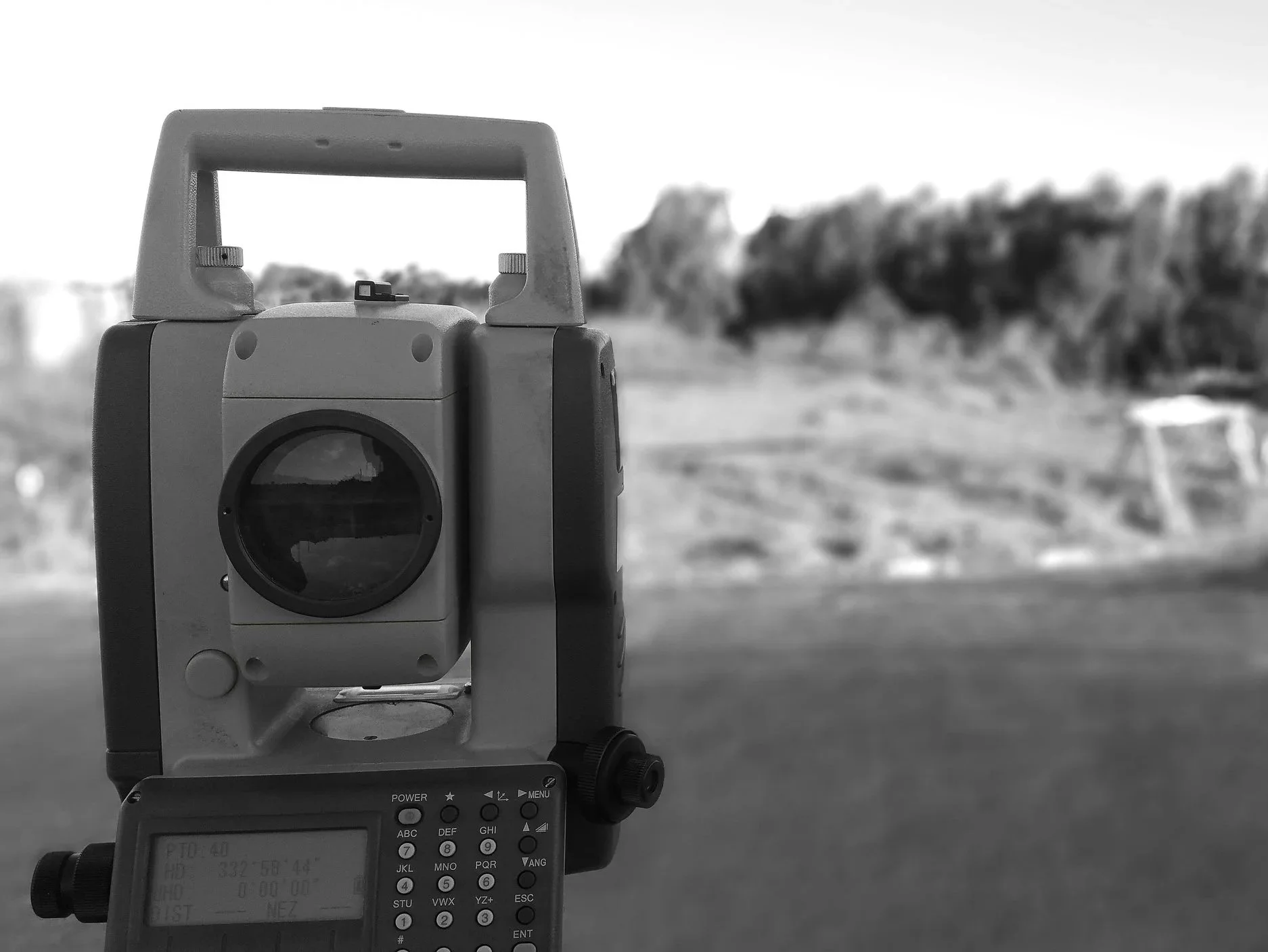 Surveyor's level measuring instrument with a focus on lens, set against a blurred outdoor background with trees and an open field.
