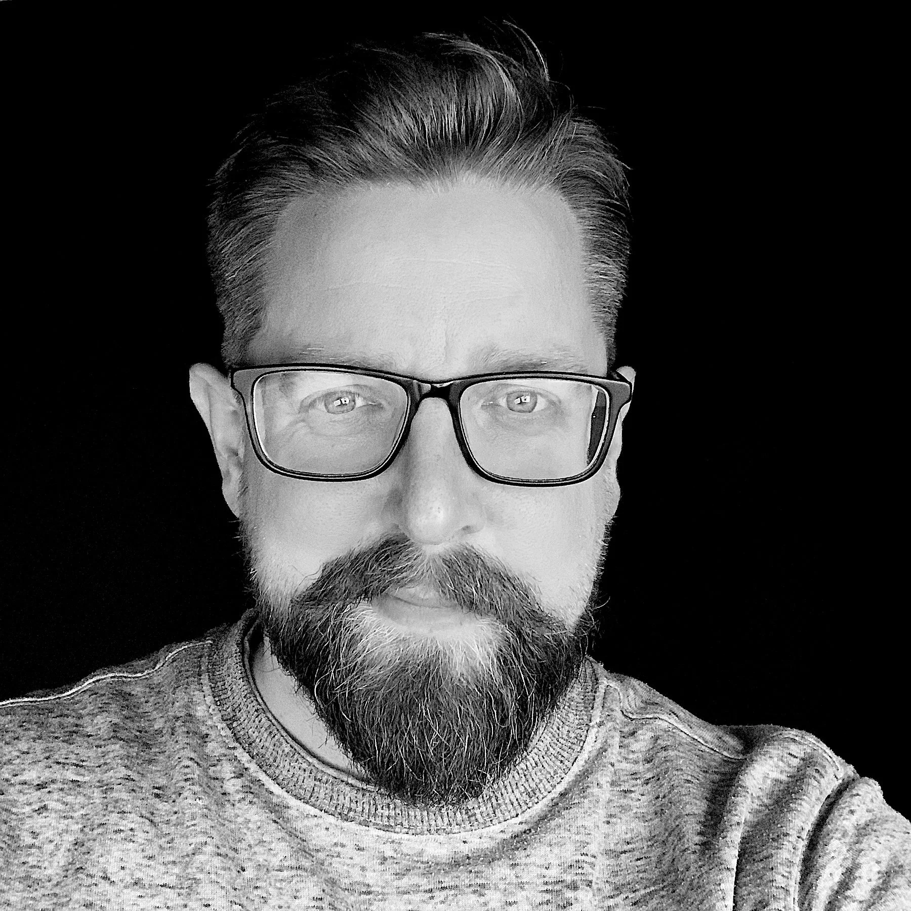 Black and white photo of a middle-aged man with glasses, a beard, and styled hair, wearing a knit sweater.